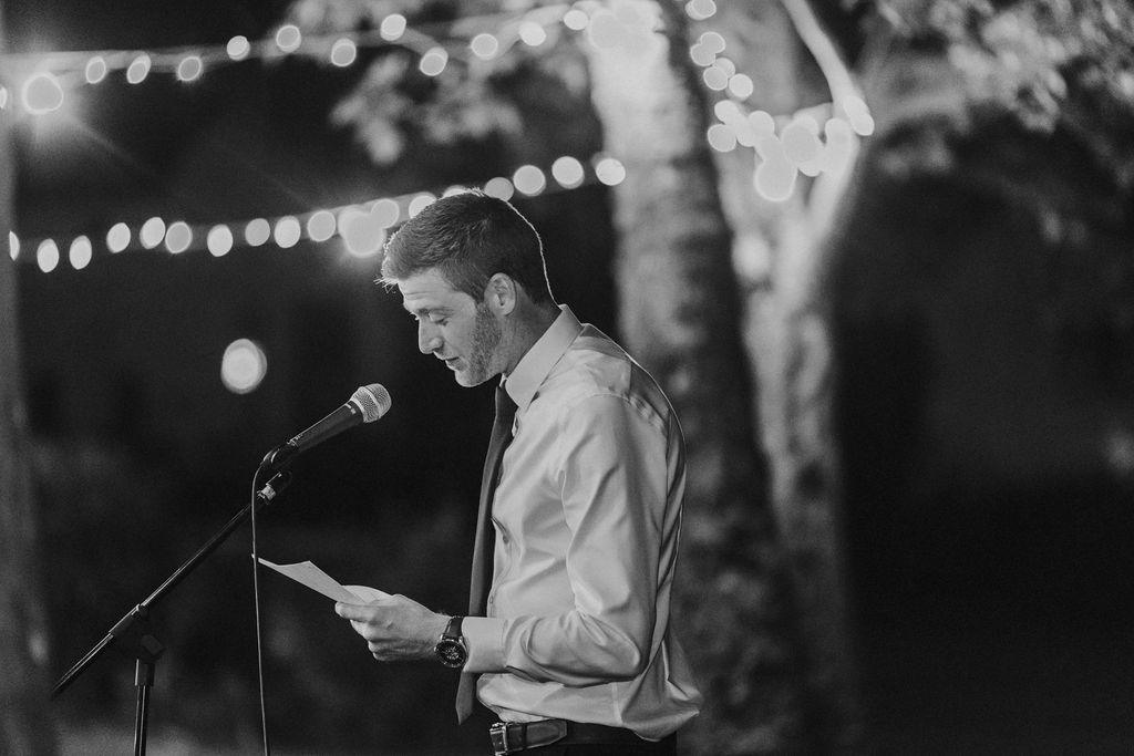 Man delivers heartfelt speech at outdoor wedding reception microphone under string lights.