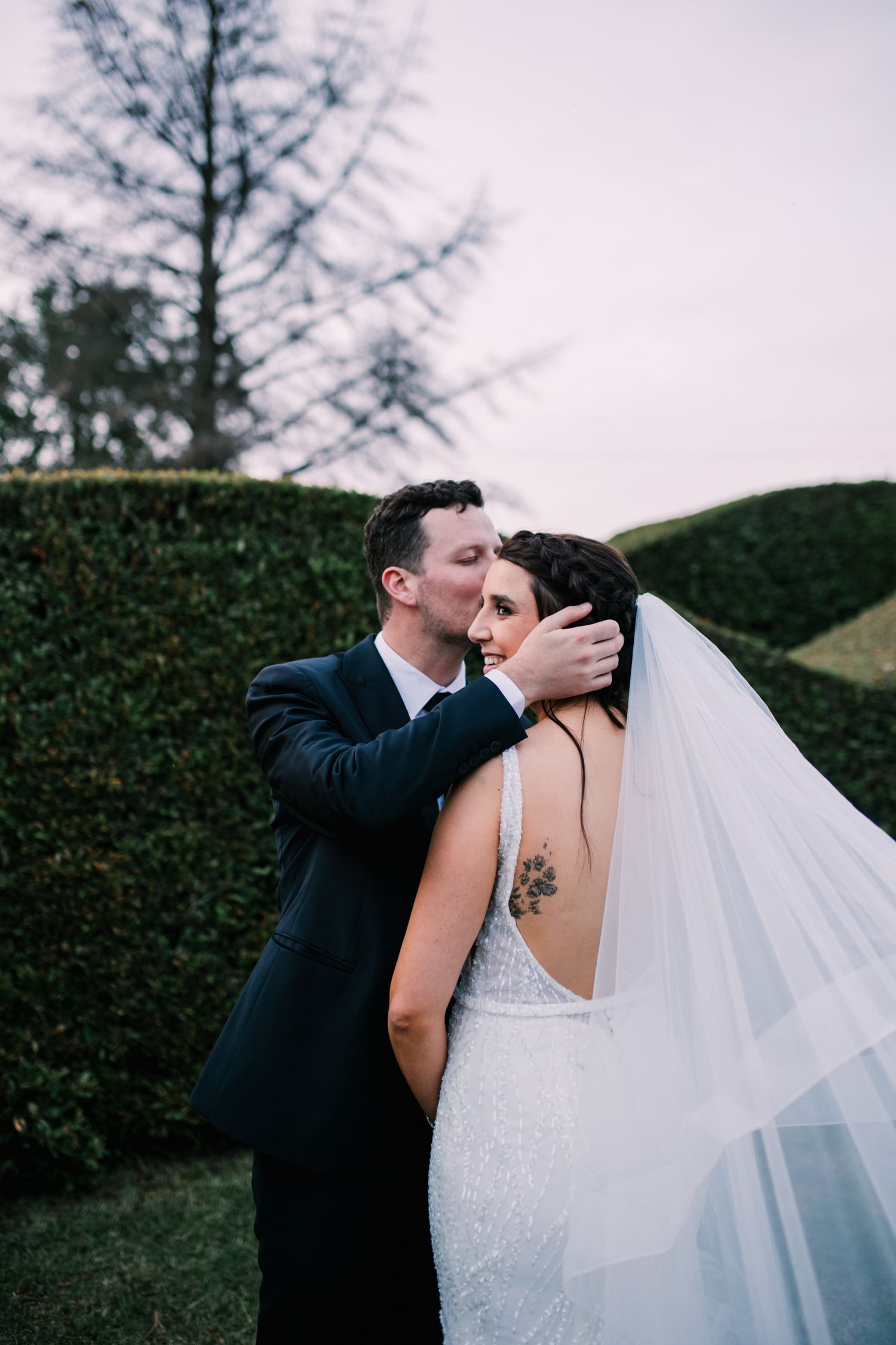 Groom kissing bride on the head while she smiles, her veil flowing softly in the breeze.
