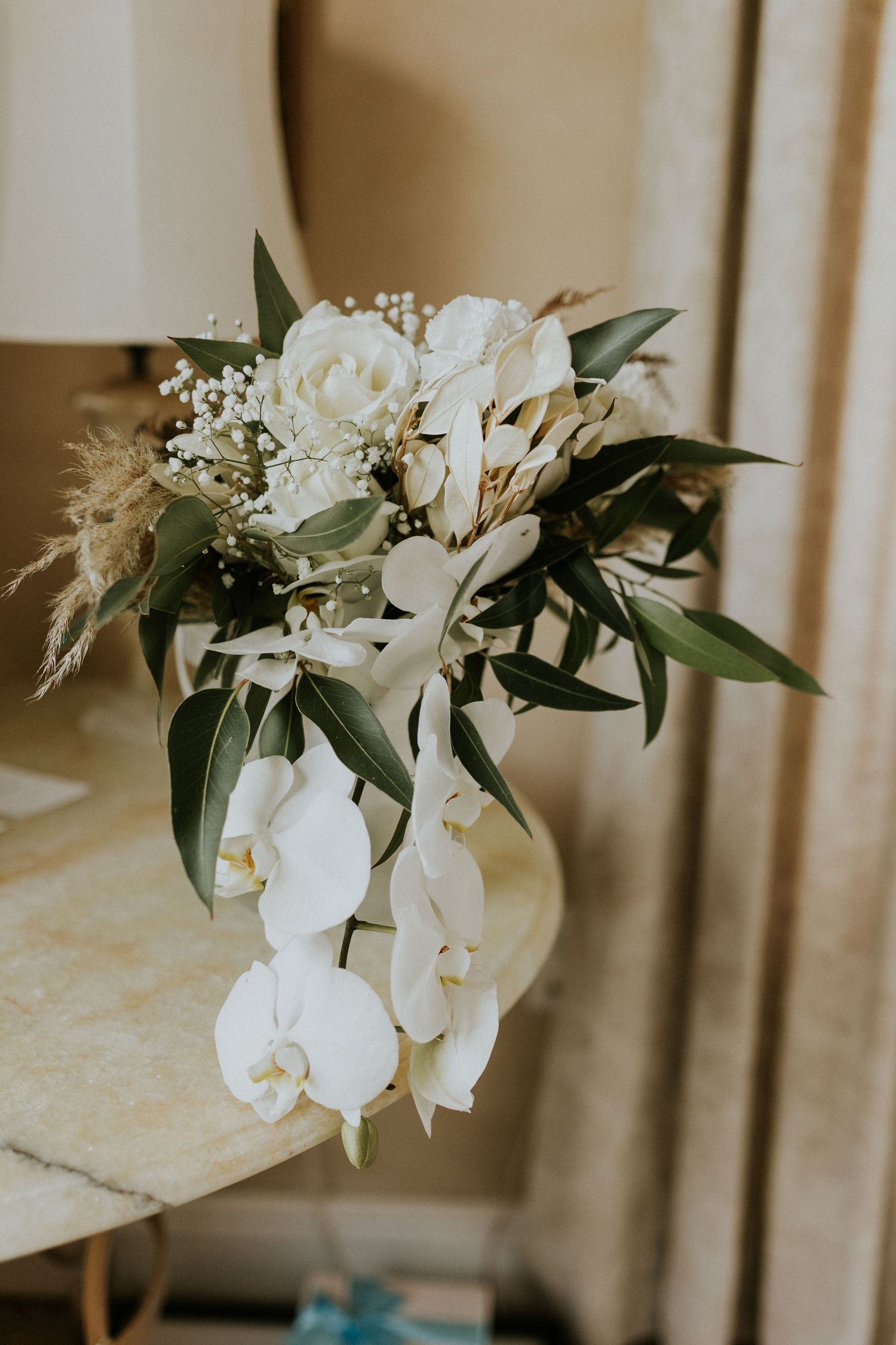 Elegant bridal bouquet featuring white orchids, roses, hydrangeas, baby's breath, and greenery on a marble side table.