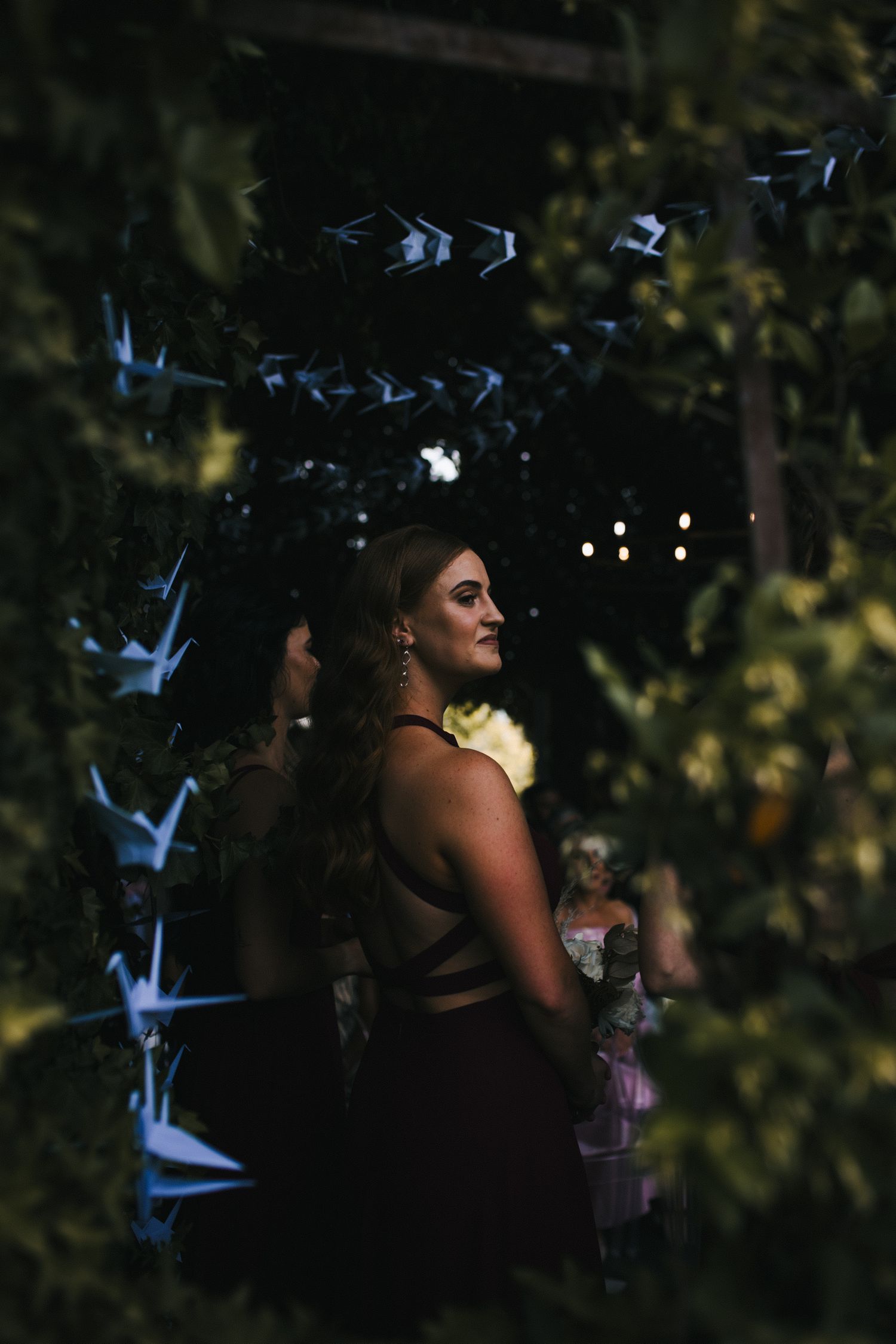 Bridesmaid in a dark backless gown stands among hanging paper cranes and string lights in a lush garden setting.