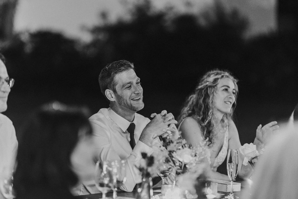 Bride and groom share a laugh together at outdoor wedding reception dinner table.