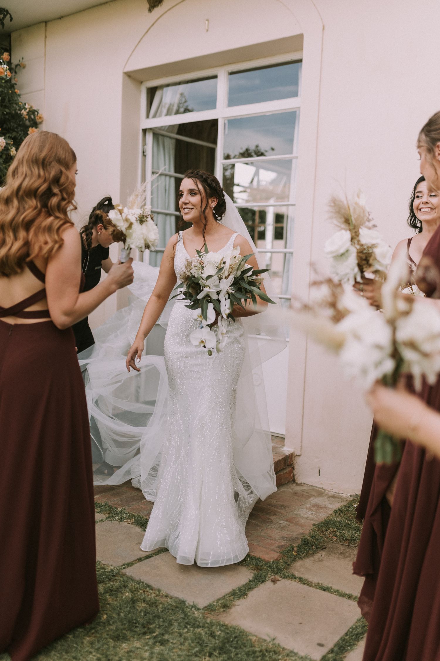 Smiling bride in a fitted lace wedding dress walks out to greet her bridesmaids in burgundy gowns holding bouquets.