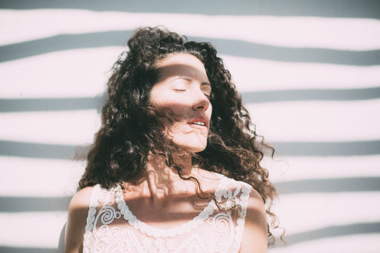 Young woman with curly hair smiling peacefully with eyes closed in striped sunlight.