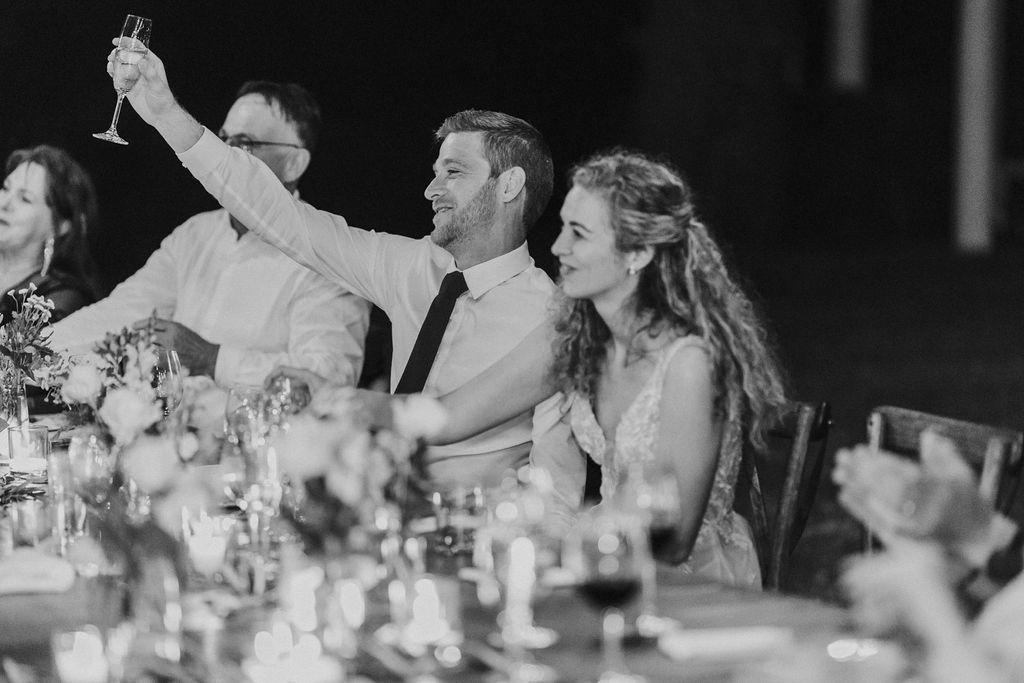Smiling couple raises champagne glass in a toast at a wedding reception dinner table.