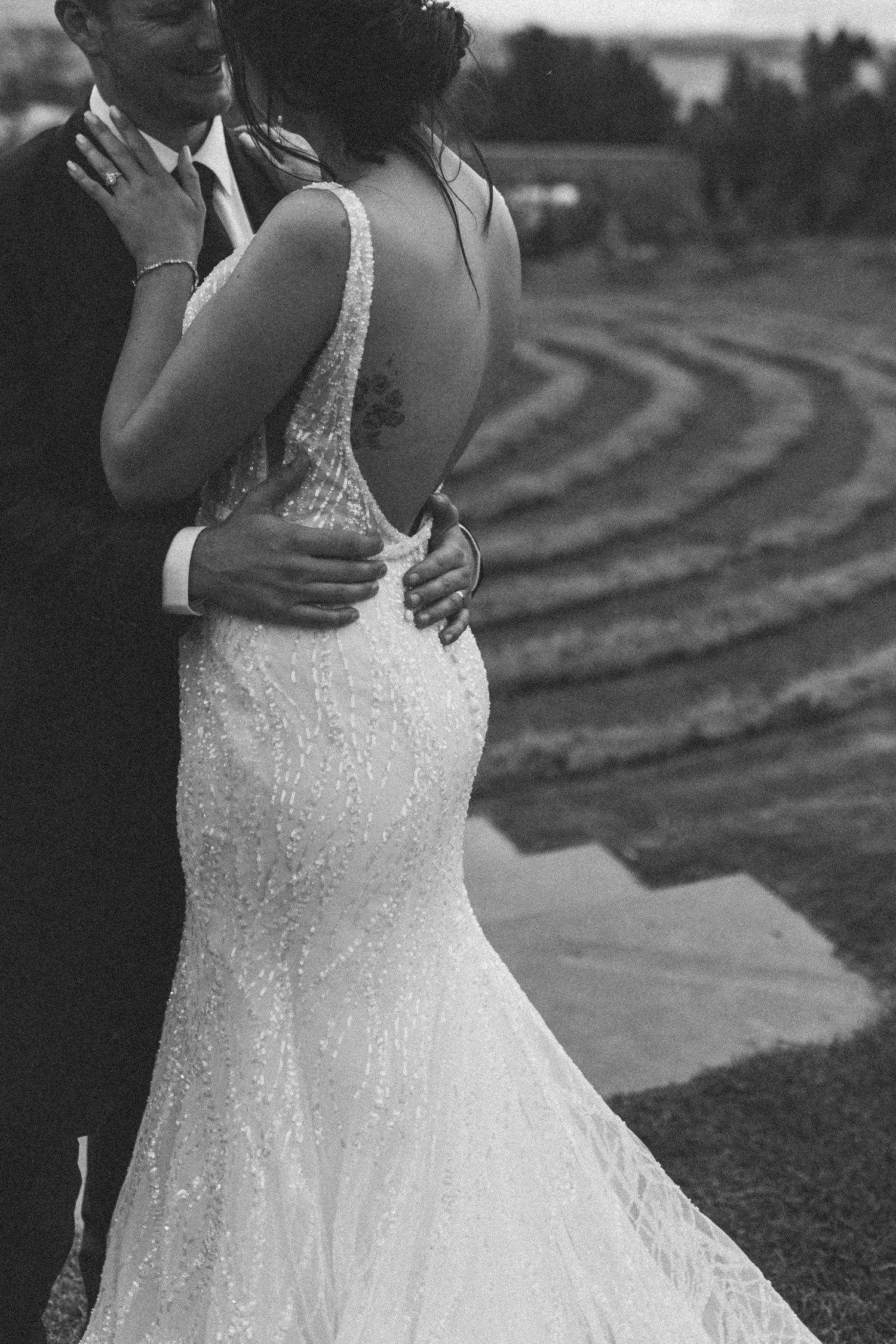 Black and white close-up of groom embracing bride in beaded backless mermaid wedding gown outdoors.