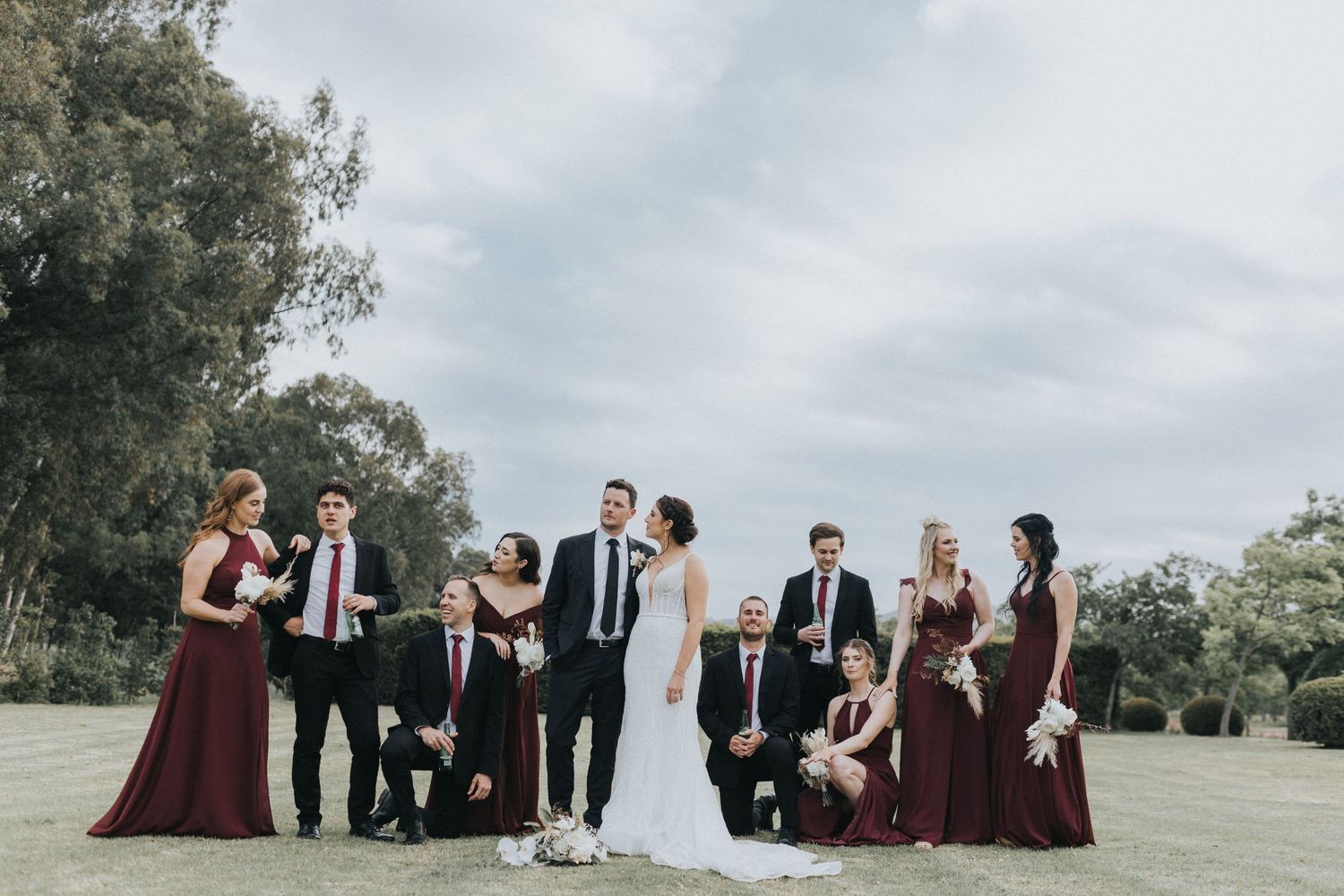 Wedding party posing outdoors in burgundy bridesmaid dresses and black suits with scenic greenery background.