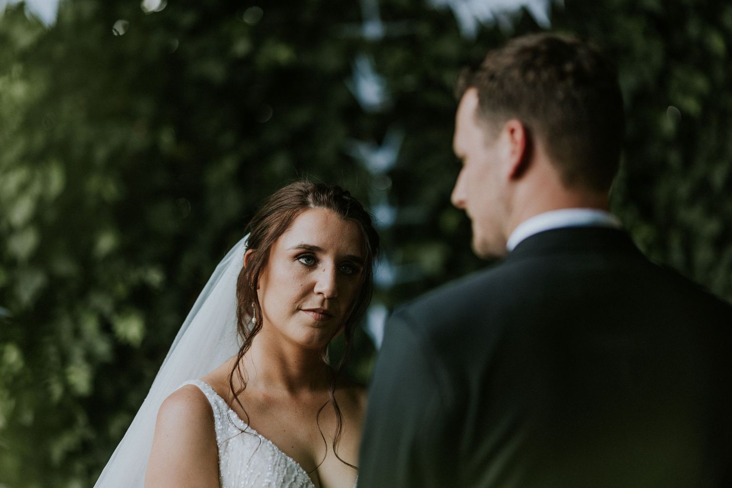 Bride in white lace gown and veil gazing at groom during emotional outdoor wedding ceremony.