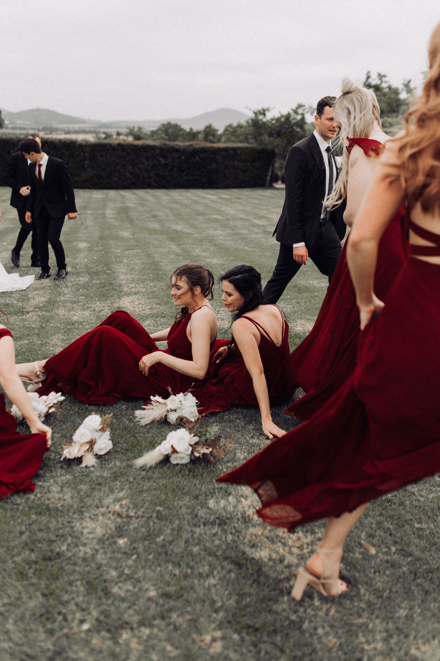 Bridesmaids in burgundy gowns laughing and sitting on grass with groomsmen at outdoor wedding reception.