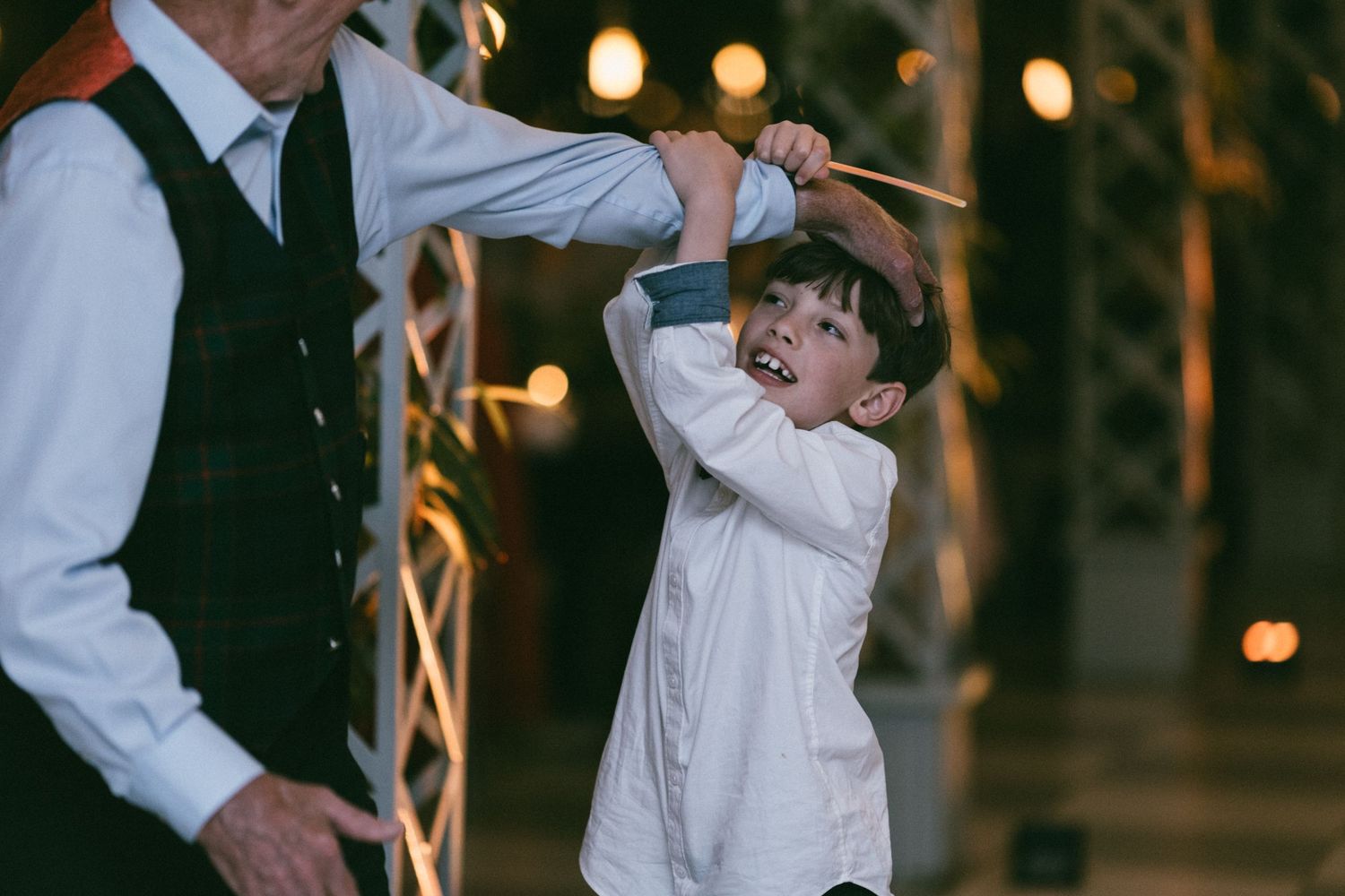 Excited young boy in white shirt playfully swinging on an adult's arm at an outdoor evening celebration.