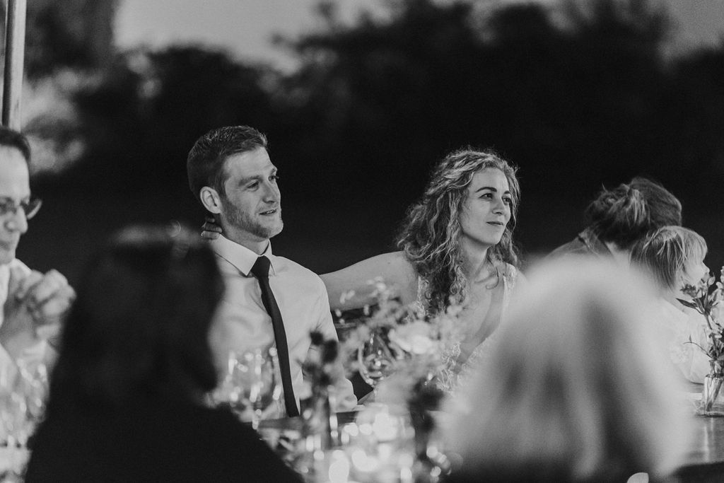 Smiling bride and groom listening to speeches at their outdoor wedding reception dinner table.