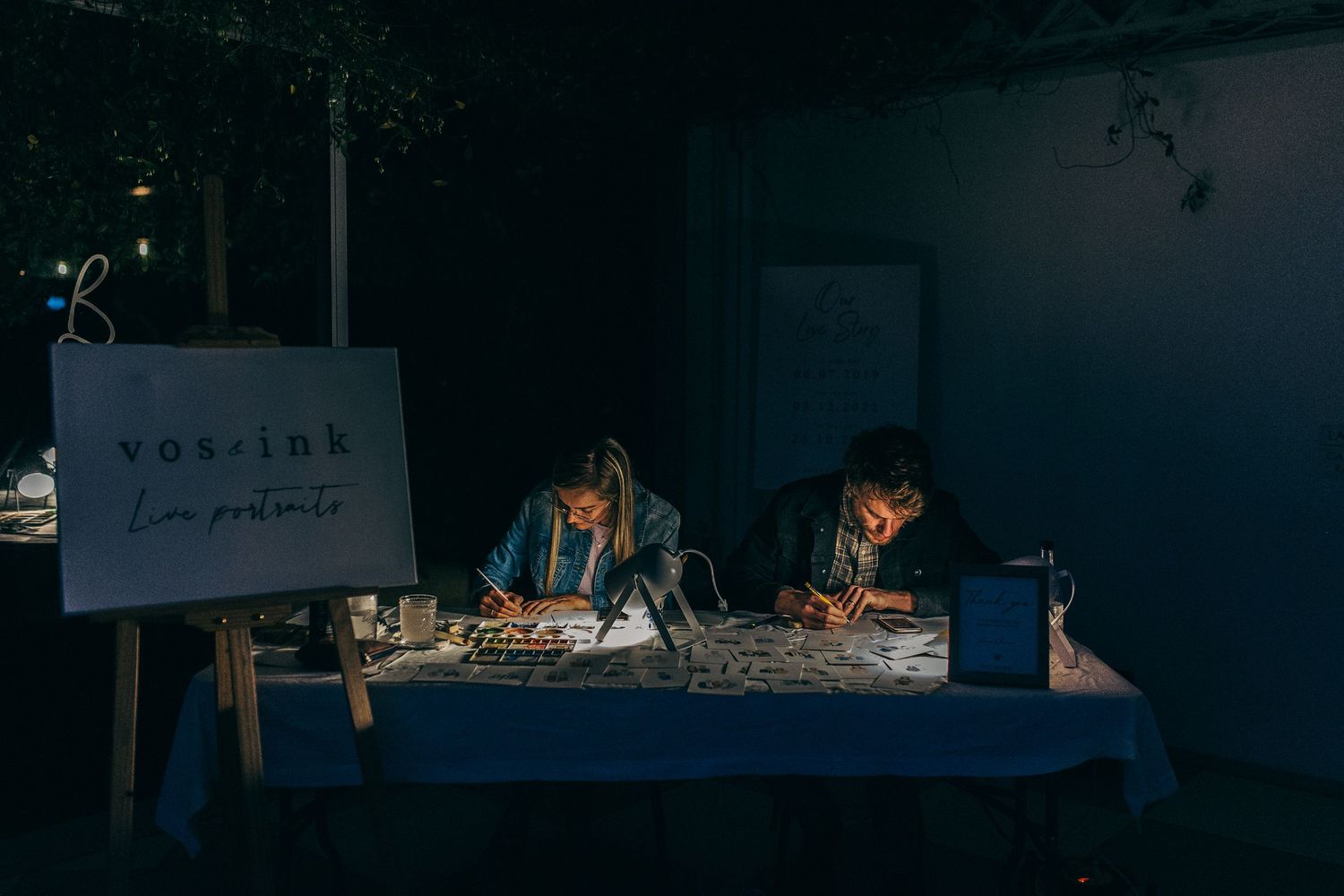 Two artists creating live portrait artwork at a dimly lit table with a Vox Ink sign at an evening event.