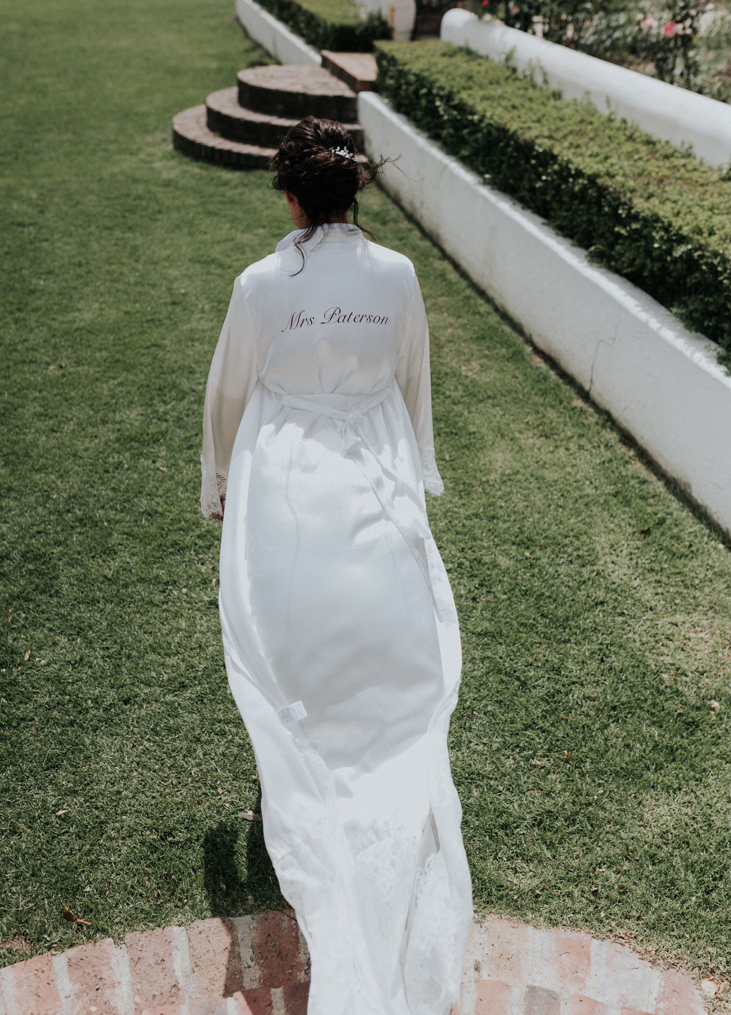 Bride in white robe with Mrs Patterson written on back walking through lush garden.