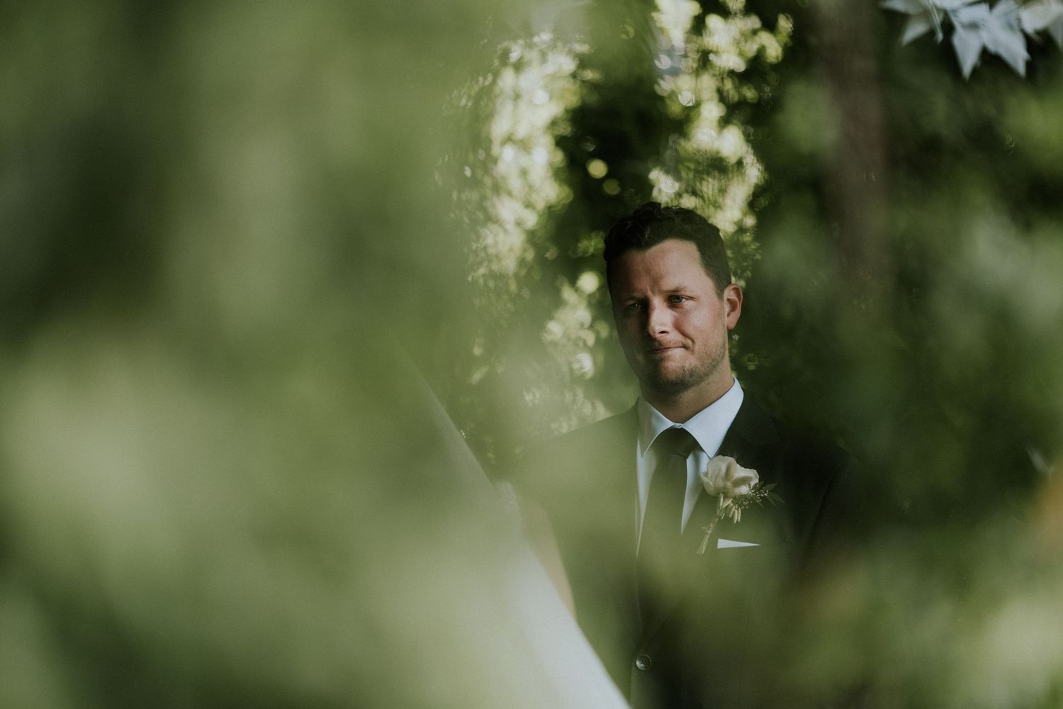Groom in grey suit with boutonniere framed by lush green foliage during outdoor wedding ceremony.