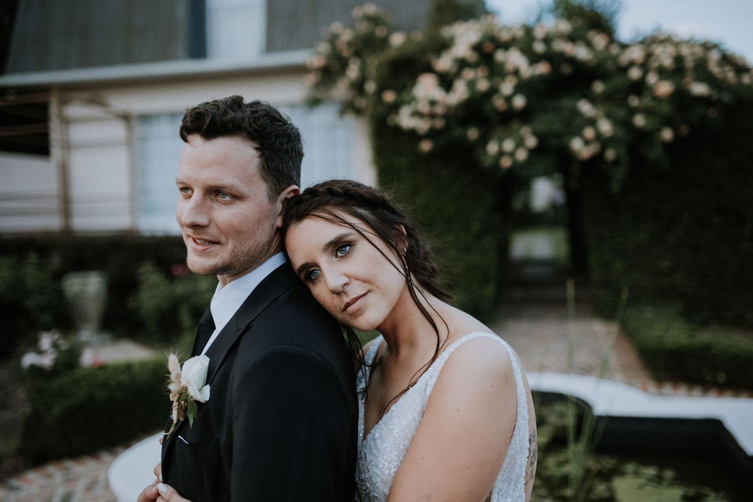 Romantic wedding portrait of bride leaning on groom's shoulder in a lush garden setting at dusk.