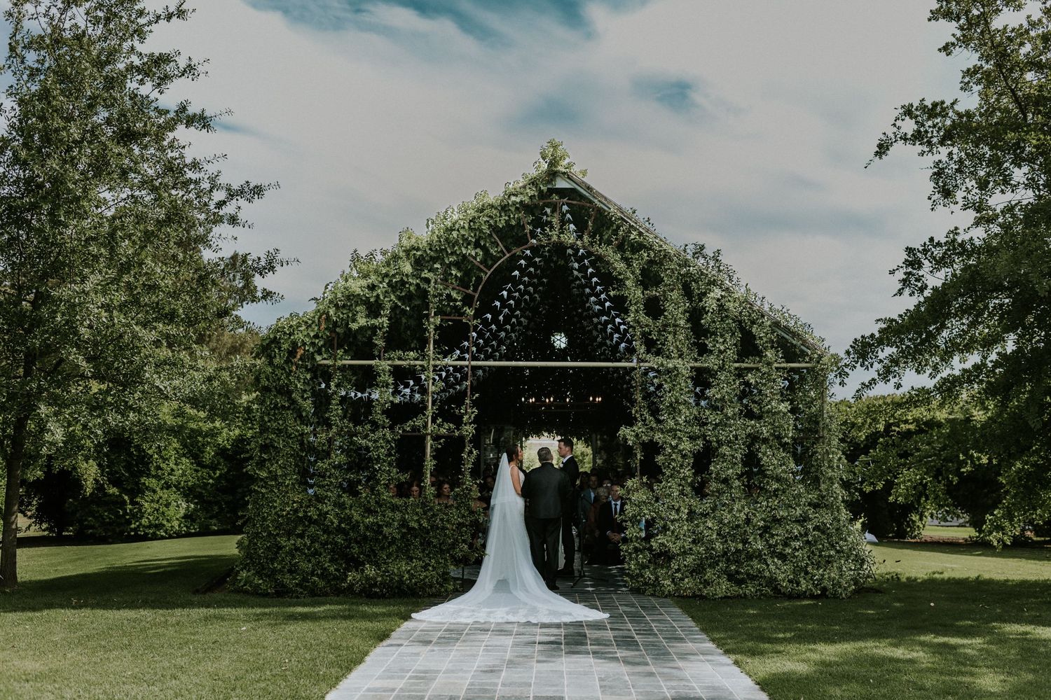 Bride and groom stand before an ivy-covered chapel arch with a long cathedral veil on a lush lawn.