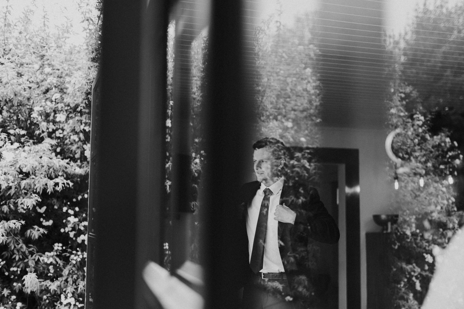 Groom adjusting tie reflected in glass doors surrounded by garden greenery.