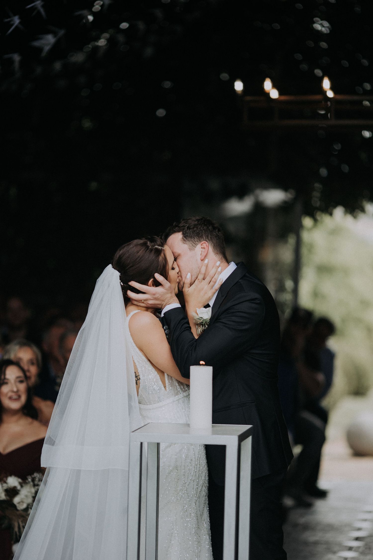 Bride and groom share first kiss at outdoor wedding ceremony with guests watching.
