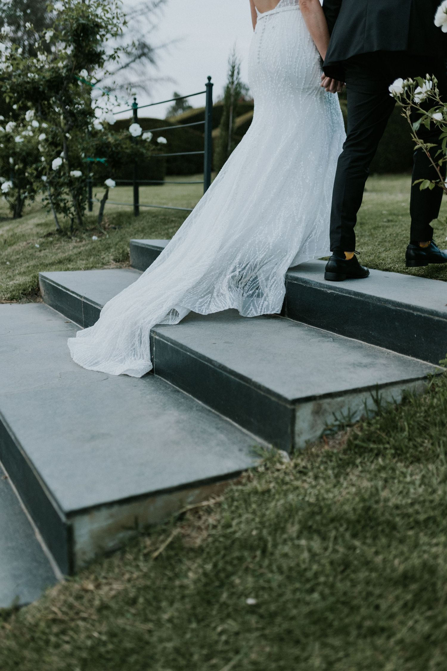 Detail shot of bride's elegant lace wedding gown train flowing down stone steps at garden venue.