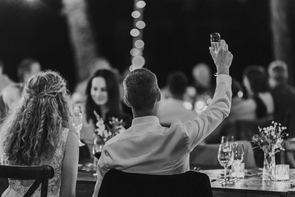 Wedding guest raises glass in toast at elegant outdoor reception with string lights in background.