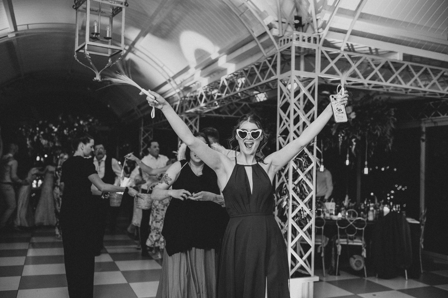 Woman in black dress with sunglasses raises arms joyfully on wedding reception dance floor at night.