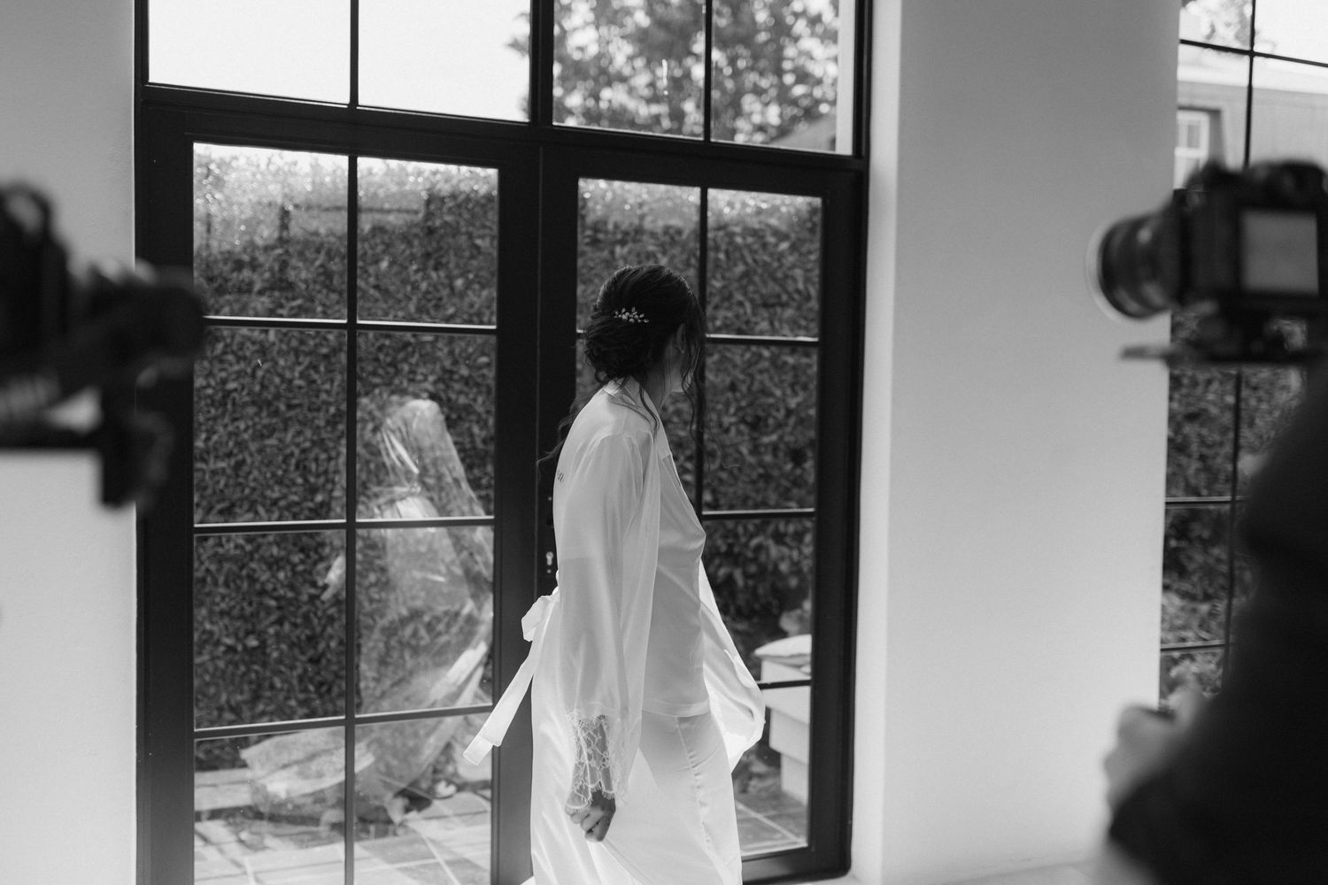 Bride in white robe stands before glass doors as photographer captures moment.