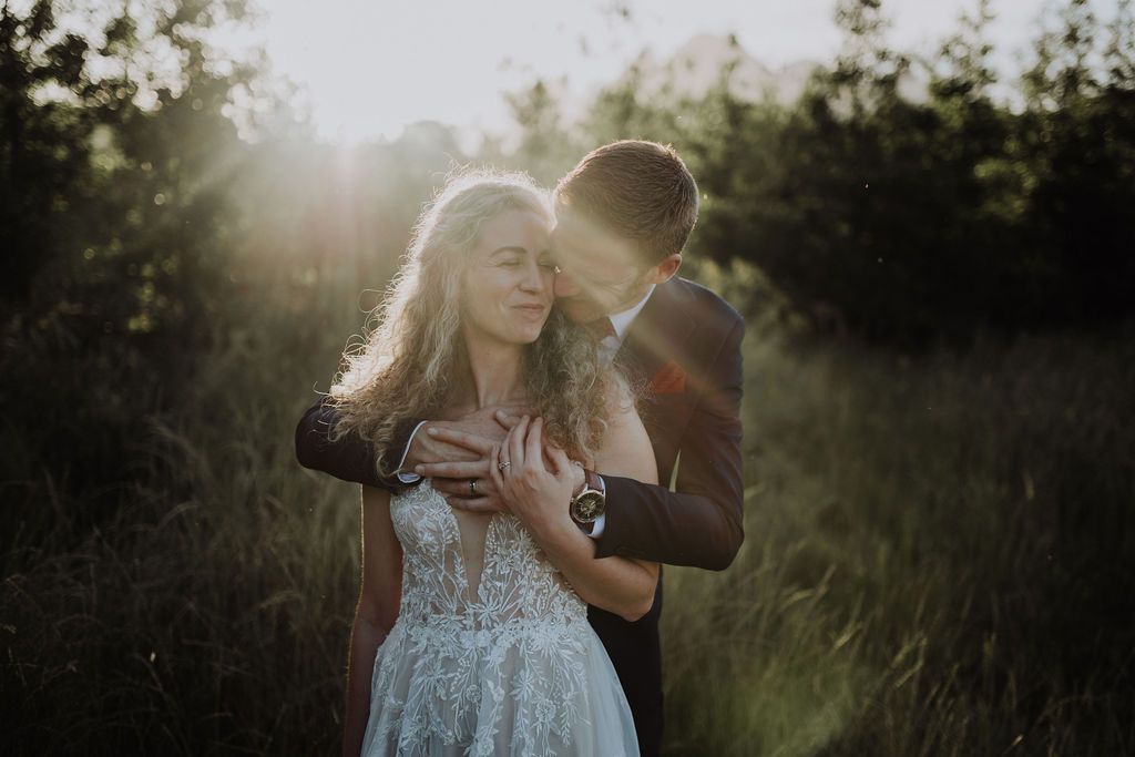 Groom embracing bride from behind in a sun-drenched field, with warm lens flare illuminating the scene.
