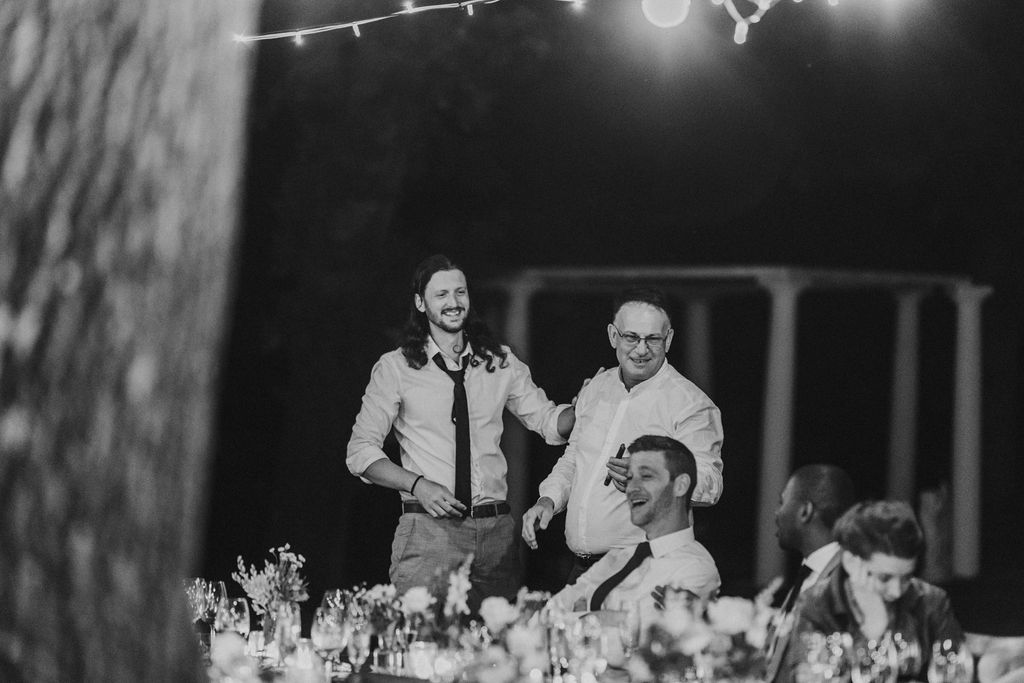 Black and white wedding reception photo of guests laughing and celebrating at an outdoor evening event with string lights.