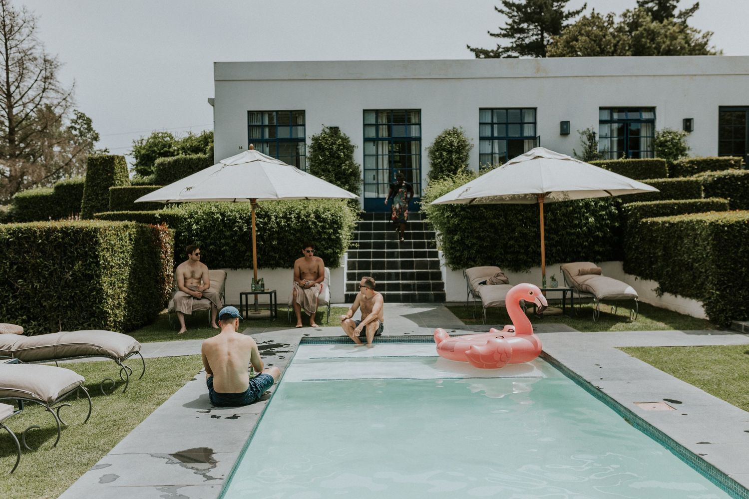 Wedding guests enjoying a sunny poolside gathering with flamingo float and white market umbrellas.