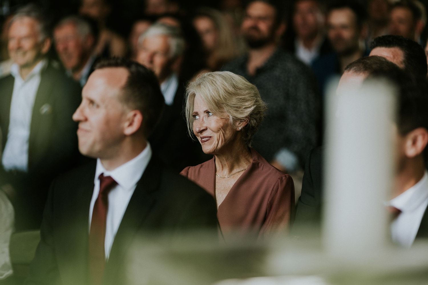 Elegant older woman smiling warmly among wedding guests during indoor ceremony.