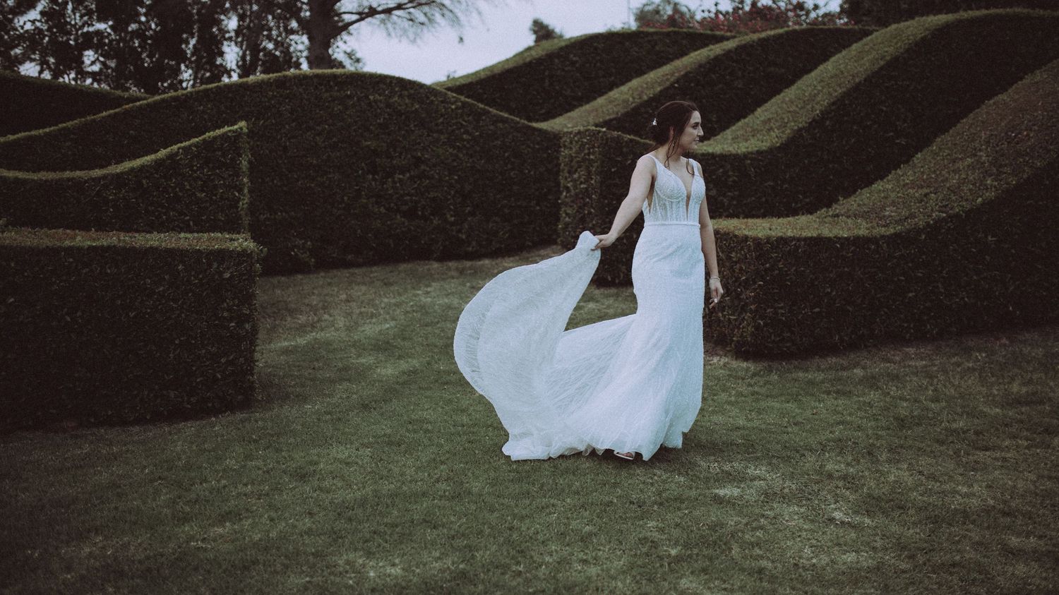 Bride twirls in flowing white gown amid sculpted wave-shaped topiary hedges in a formal garden setting.