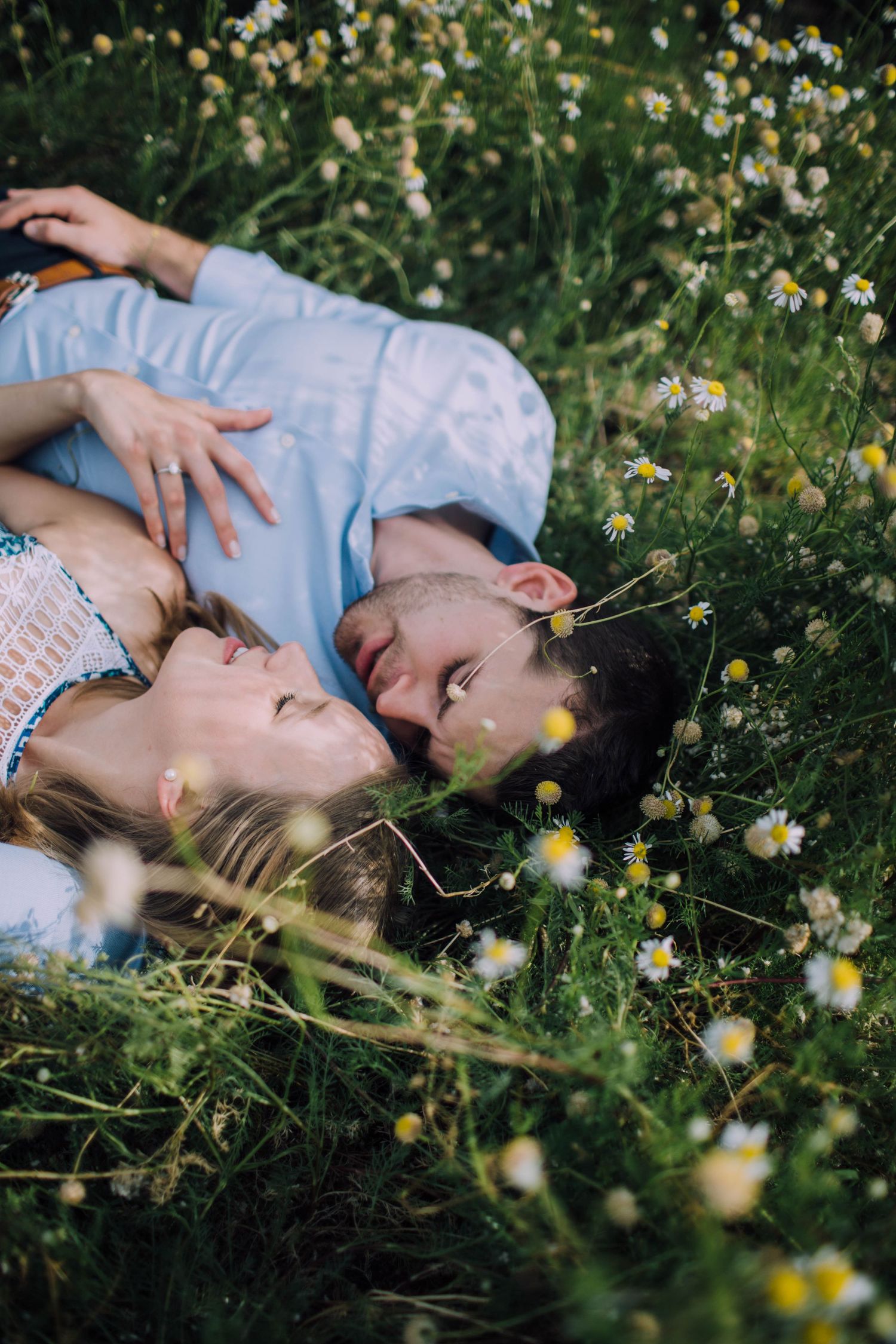 A couple lies together in a field of chamomile flowers with eyes closed in peaceful embrace.