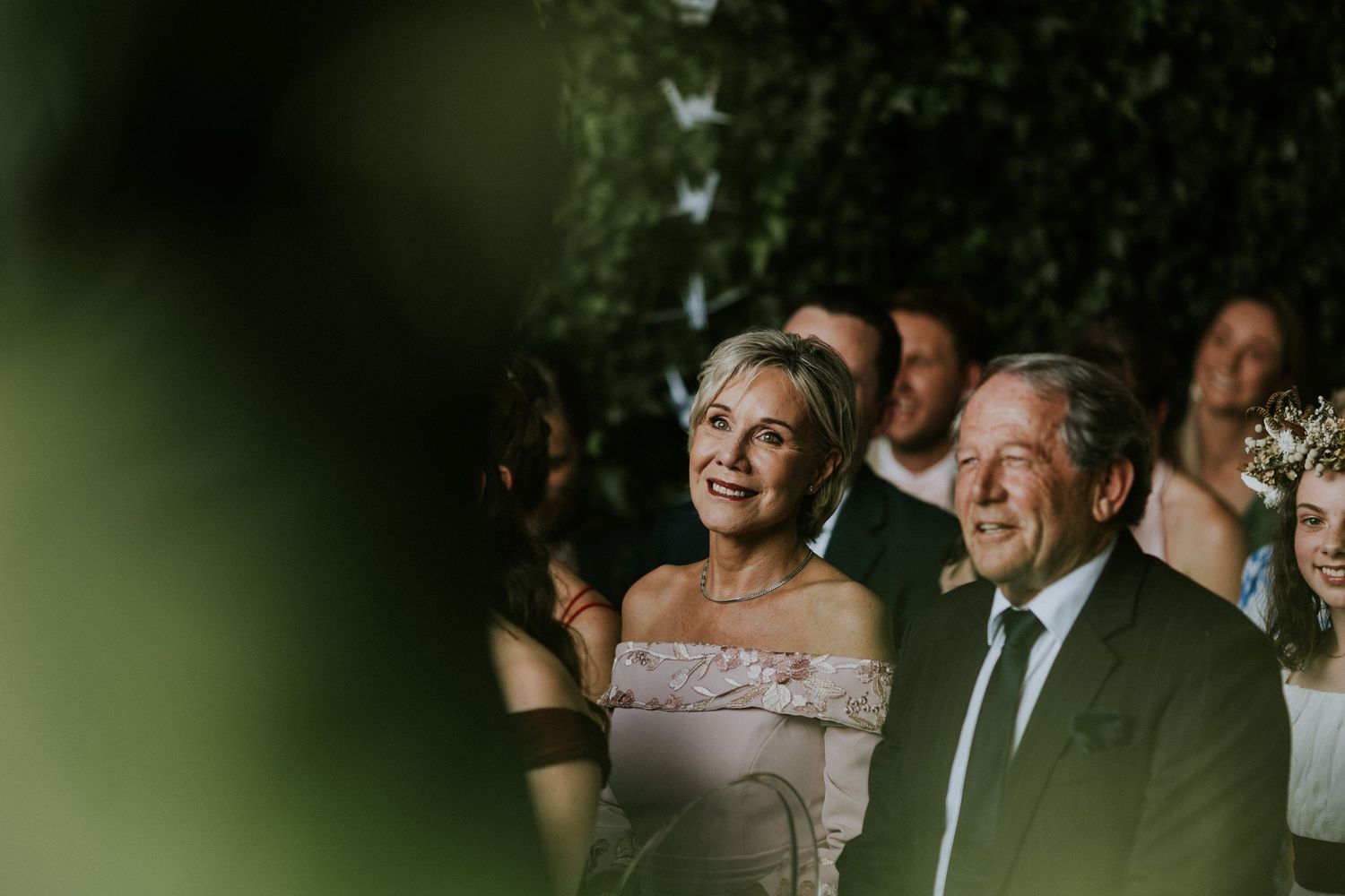 Smiling wedding guests, an elegant woman in off-shoulder dress and older man in suit, watching ceremony outdoors.
