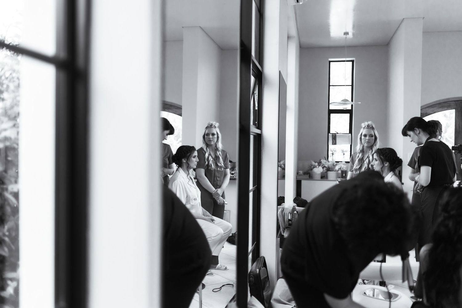 Black and white photo of bridal party getting hair and makeup done in a bright salon mirror.