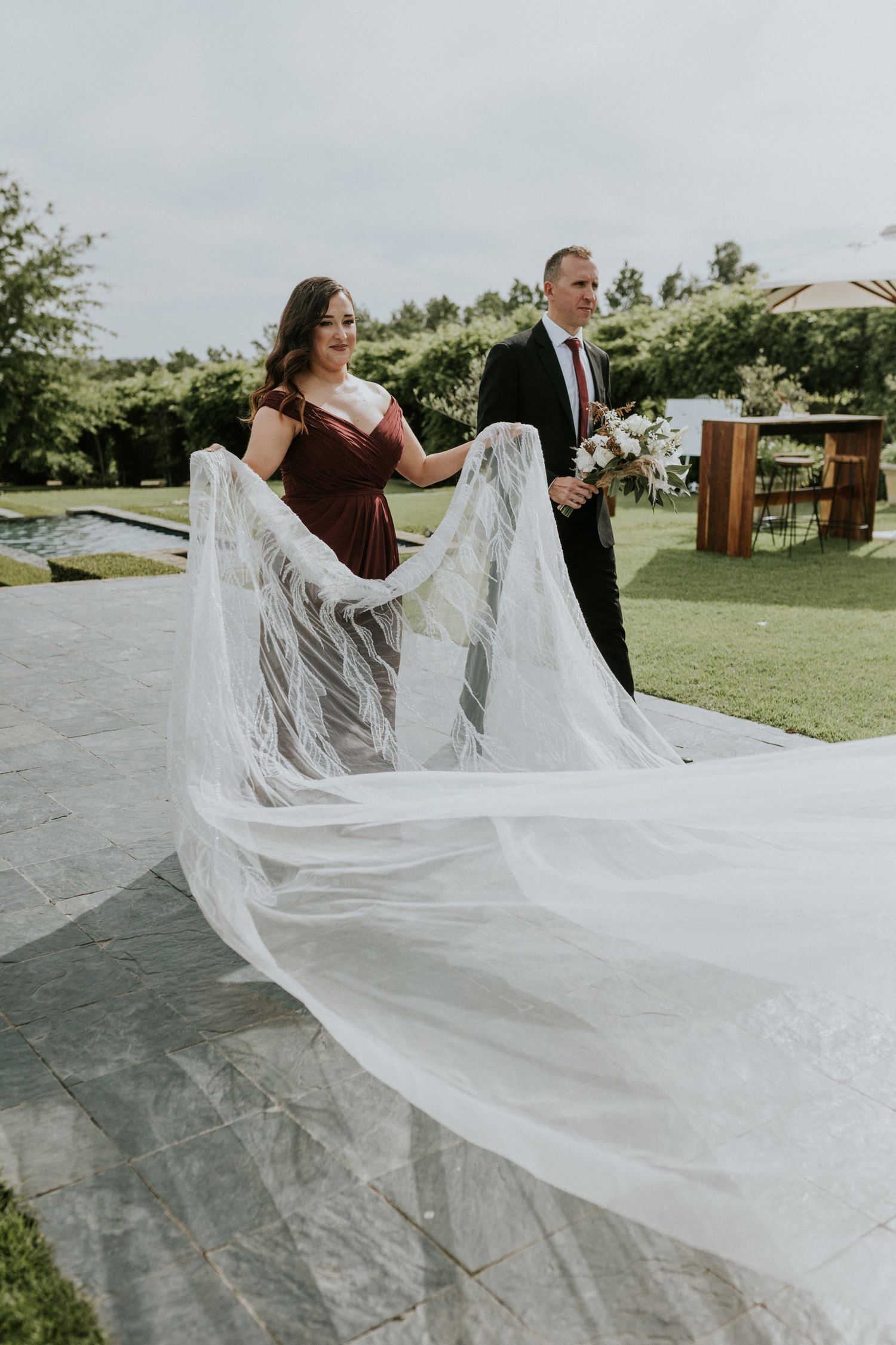Bridesmaid in burgundy dress holds the bride's flowing white veil outdoors at a garden wedding venue.