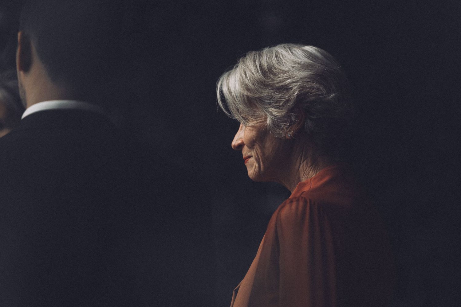 Elegant older woman in red cape smiling softly in profile at a wedding celebration.