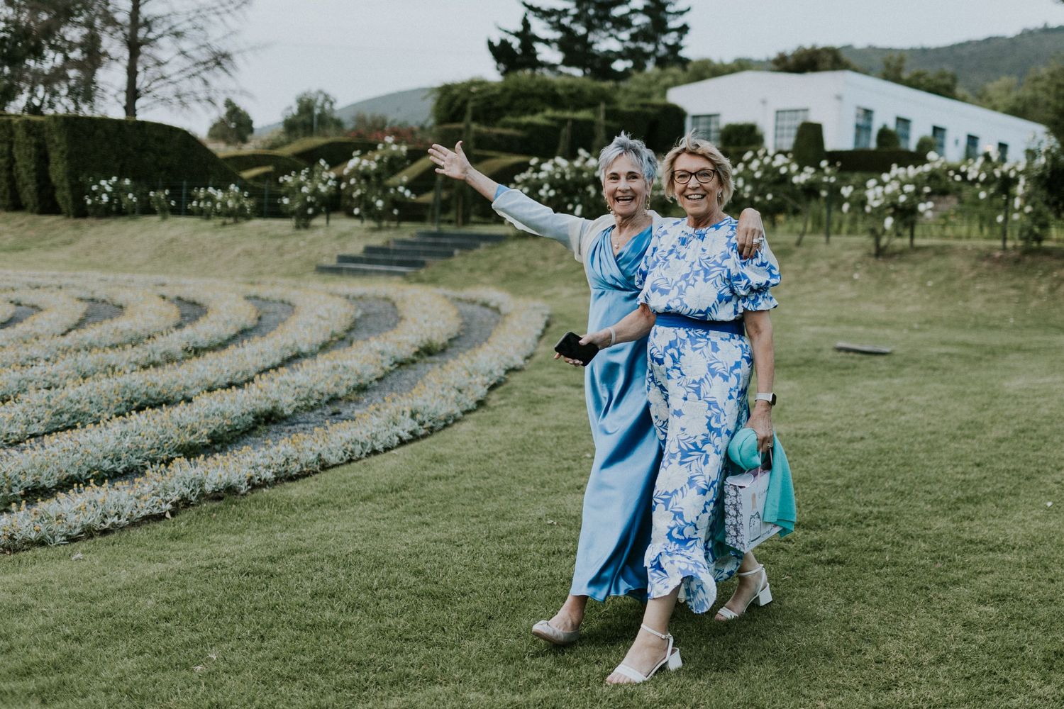 Two elegantly dressed women in blue floral outfits posing and smiling in a manicured garden.
