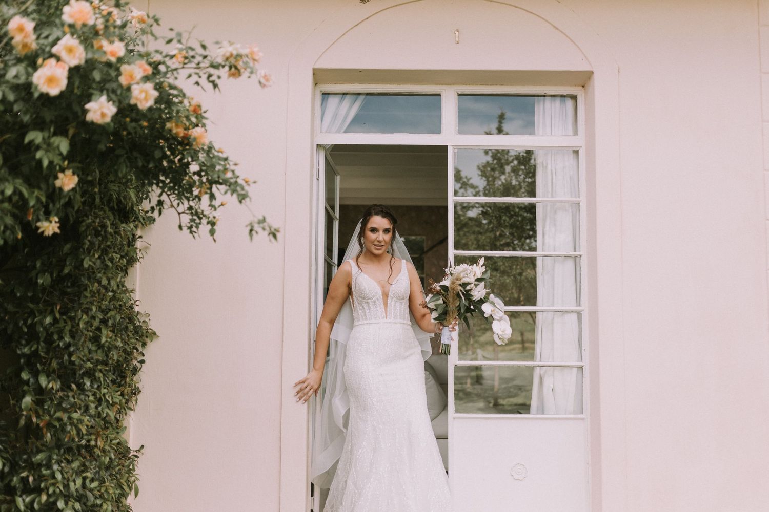 Elegant bride in a lace wedding gown holding a bouquet stands in a doorway of a pink building with climbing roses.
