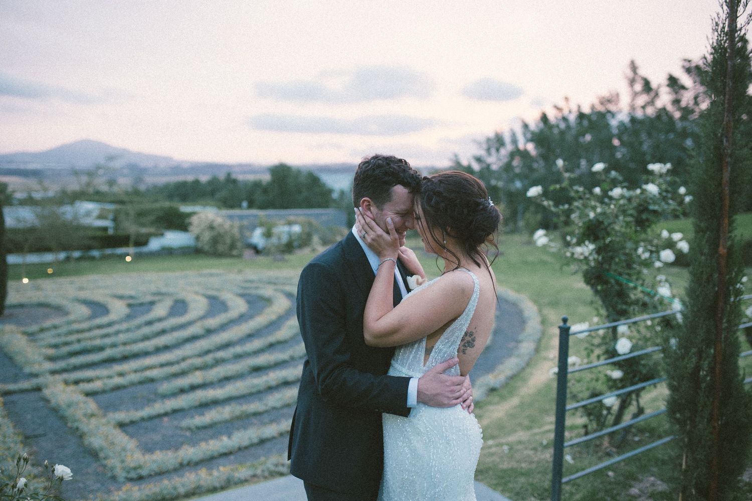 Couple shares a tender kiss at sunset overlooking a scenic garden labyrinth and mountain landscape.