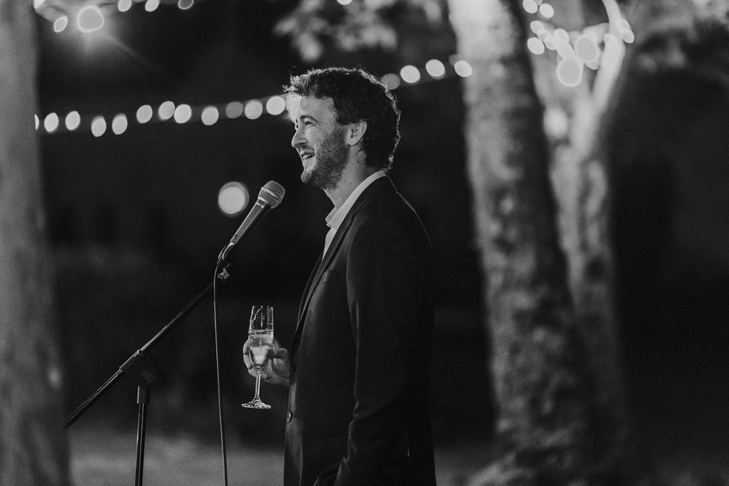 Man delivers wedding speech at microphone holding champagne glass under string lights.