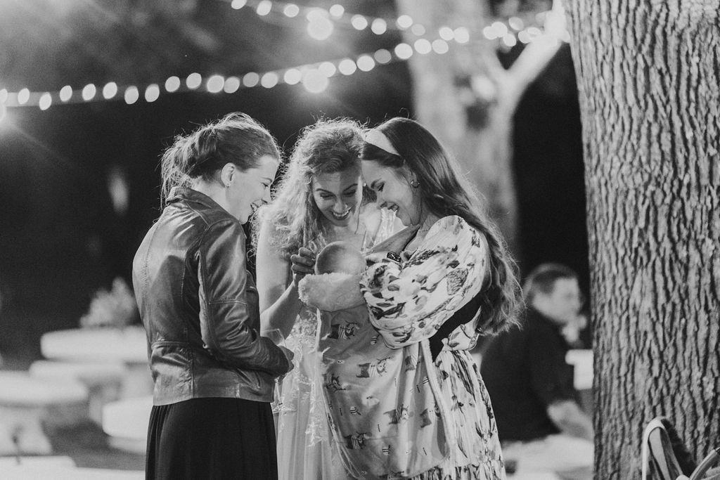 Black and white photo of three women gathered together admiring a baby at an outdoor wedding.
