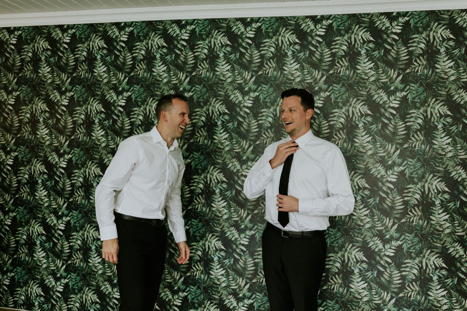 Two groomsmen in white shirts laugh together against a tropical leaf backdrop.