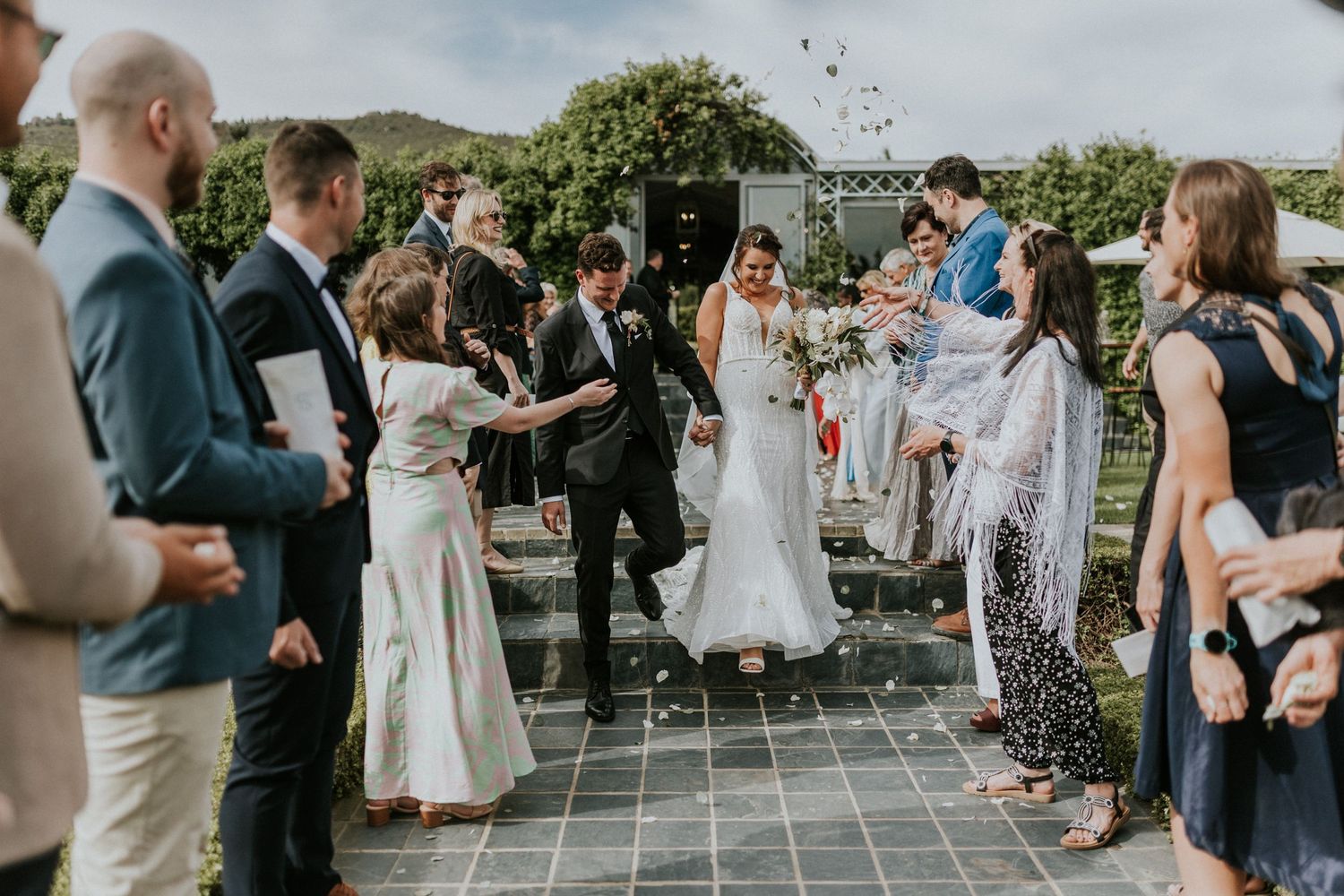 Bride and groom walk through a joyful confetti shower as wedding guests celebrate along a garden pathway.