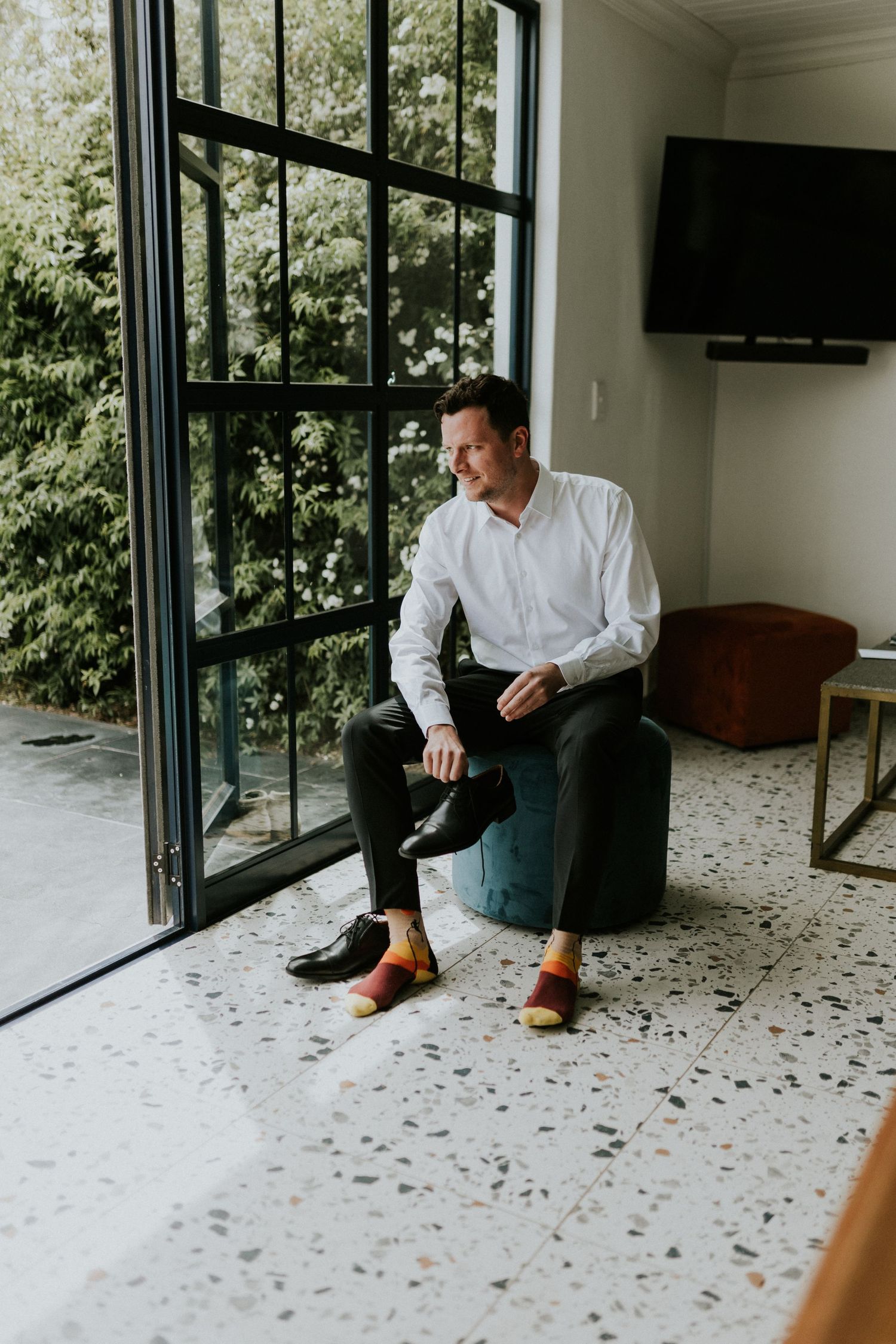 Groom in white shirt and colorful socks sits relaxed by large window before wedding ceremony.