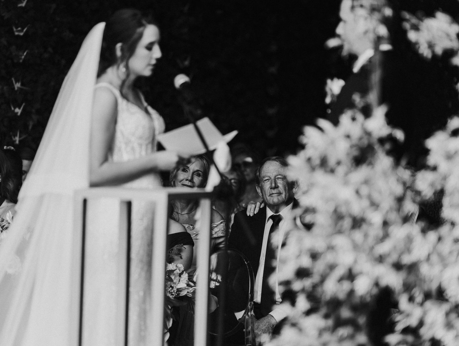 Black and white image of a bride reading her wedding vows while guests watch emotionally.