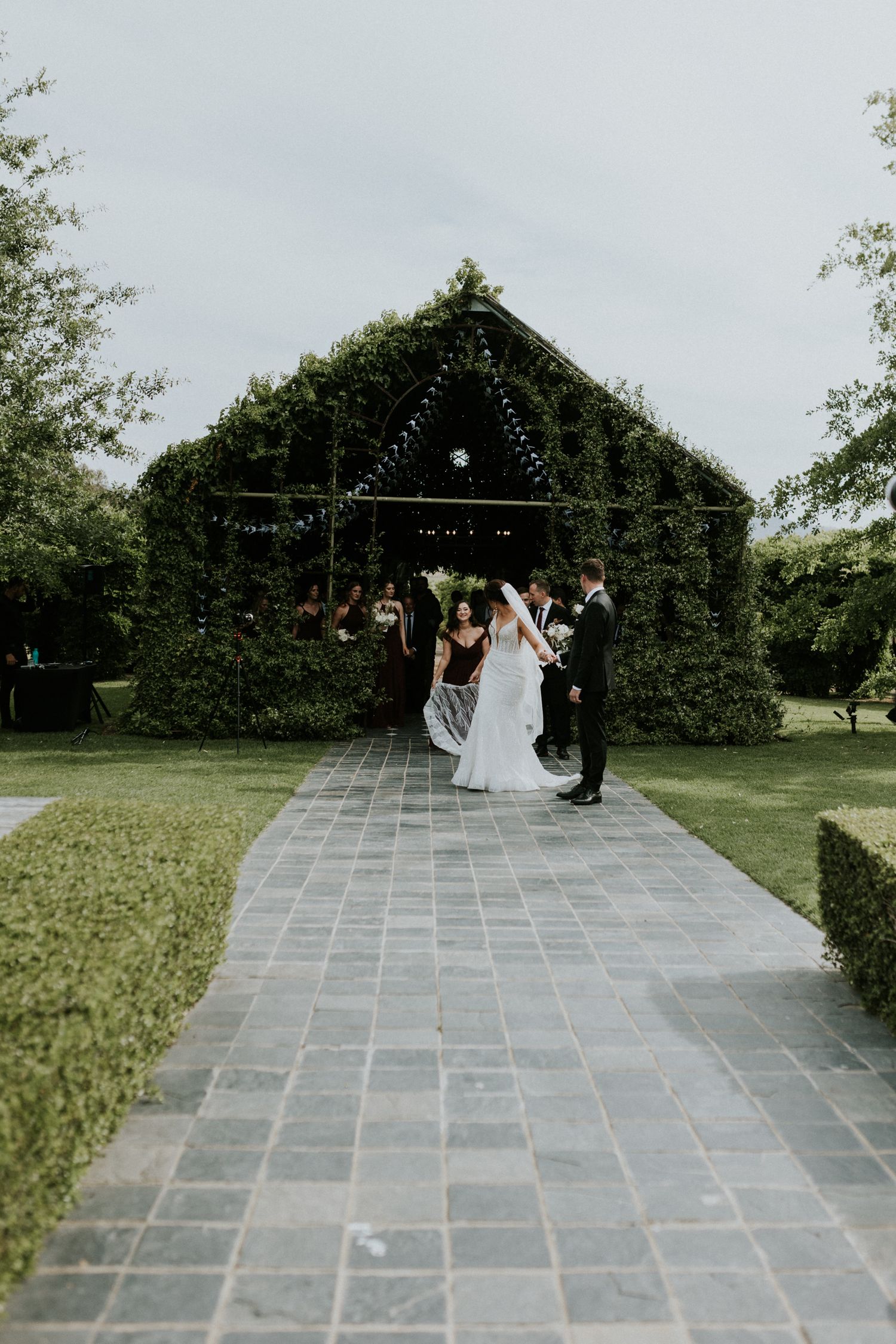 Newlyweds walk down stone pathway after ceremony at ivy-covered outdoor wedding venue.