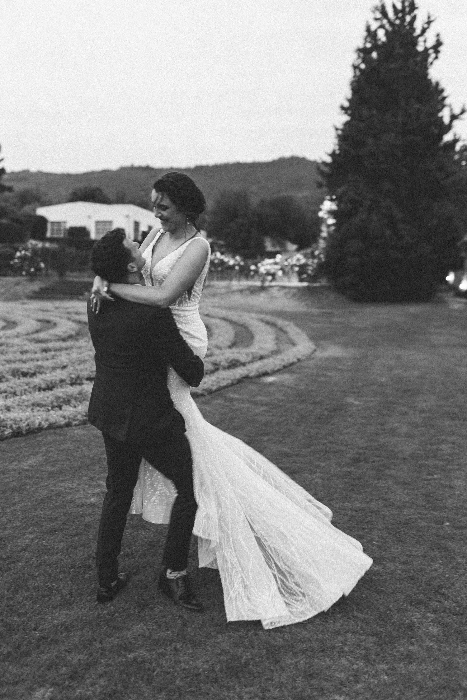 Groom lifts smiling bride in elegant lace wedding dress in joyful black and white garden photo.