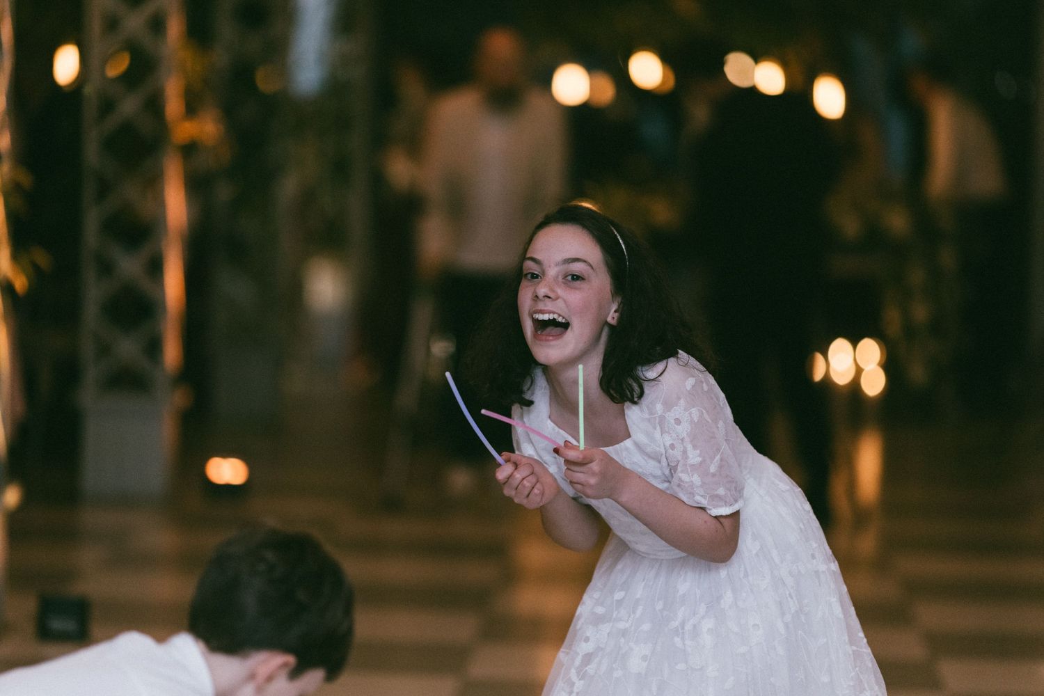 Joyful bride in white dress laughing and holding scissors at a lively wedding reception.