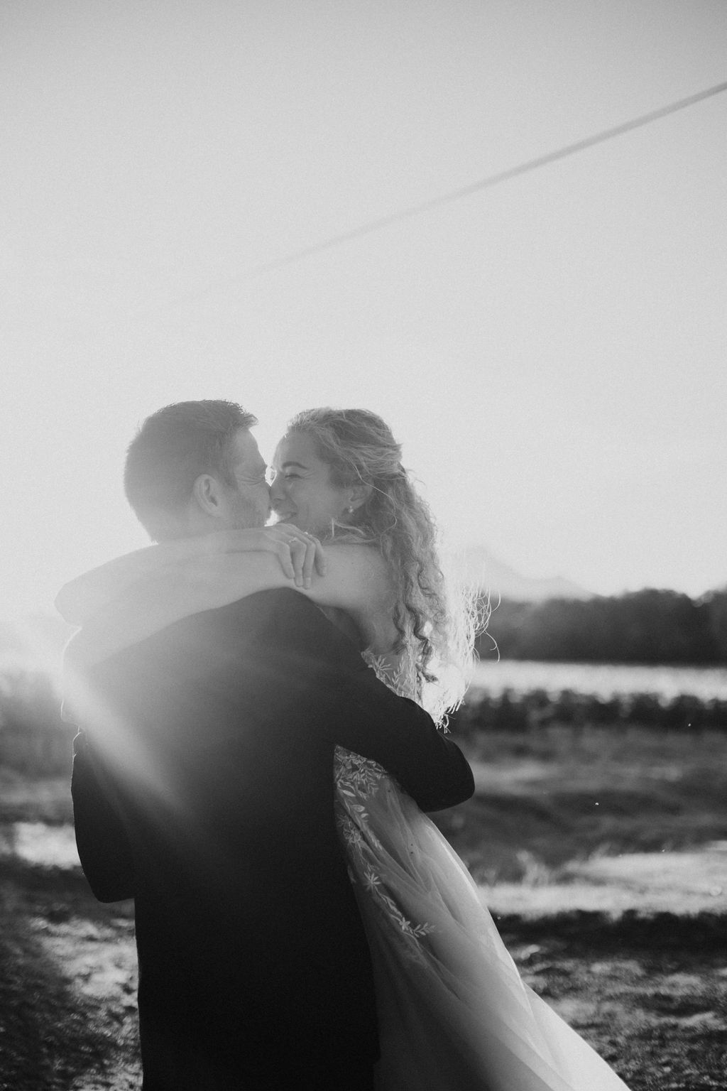 Black and white romantic wedding photo of couple kissing at sunset with lens flare effect.