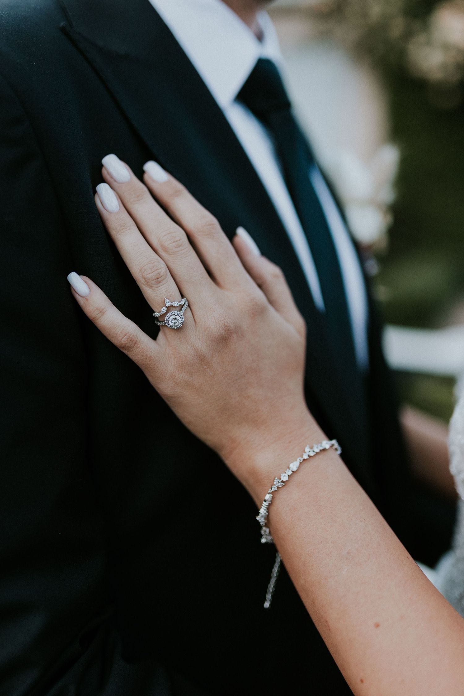Close-up of bride's hand displaying stunning diamond engagement ring and tennis bracelet on groom's suit.