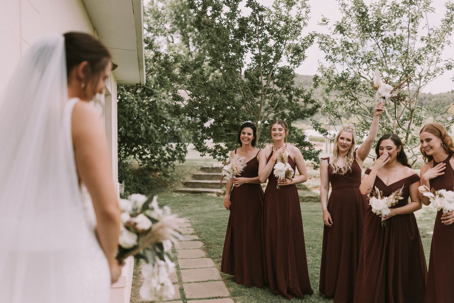 Excited bridesmaids in dark brown gowns react joyfully upon seeing the bride during a first look moment outdoors.